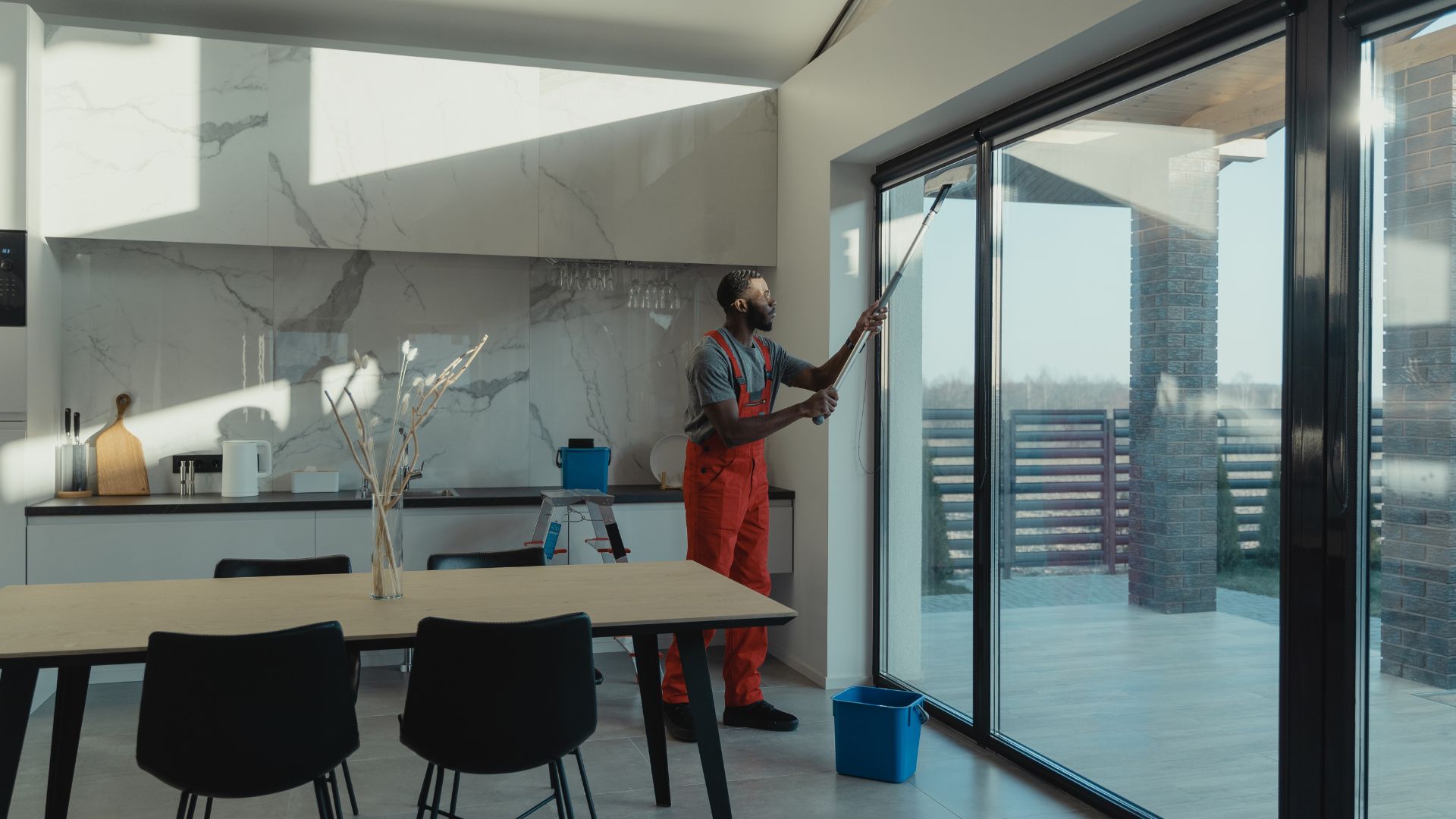 Window cleaner in red overalls washing large glass doors in modern kitchen
